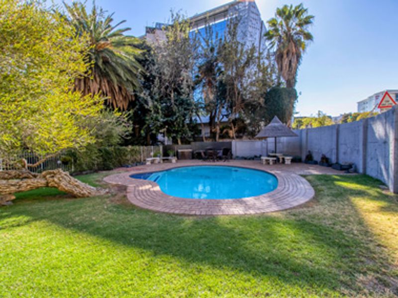 A private outdoor pool area featuring a paved surround, established garden, and a thatched umbrella, set within a secure residential complex.