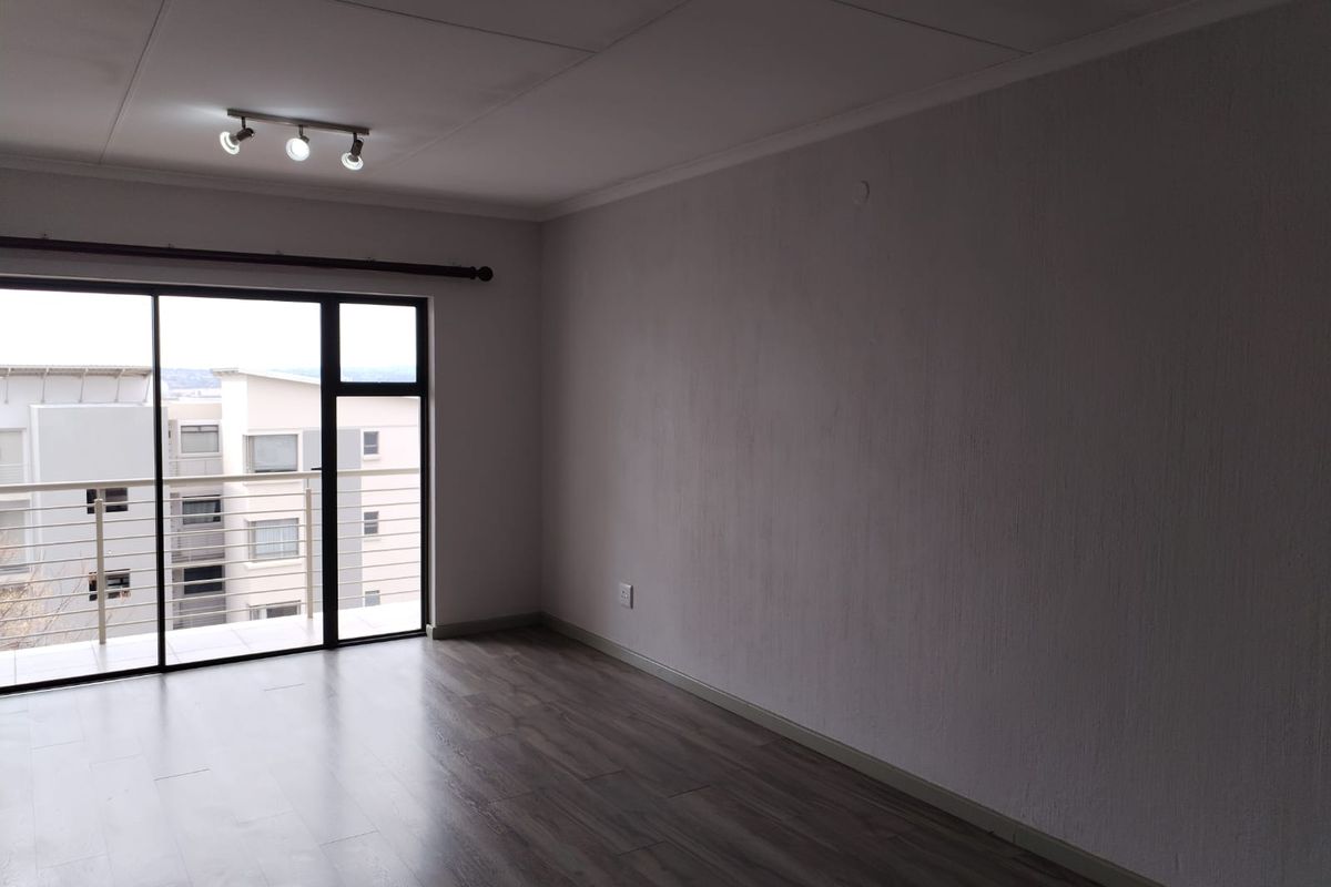A bright, empty living room featuring modern laminate flooring, track lighting, and a large sliding glass door providing access to a balcony.