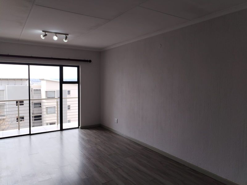 A bright, empty living room featuring modern laminate flooring, track lighting, and a large sliding glass door providing access to a balcony.