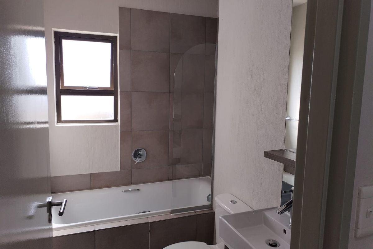 A clean, modern bathroom featuring a bathtub with a glass shower screen, a wall-mounted basin, and neutral-toned wall tiling.