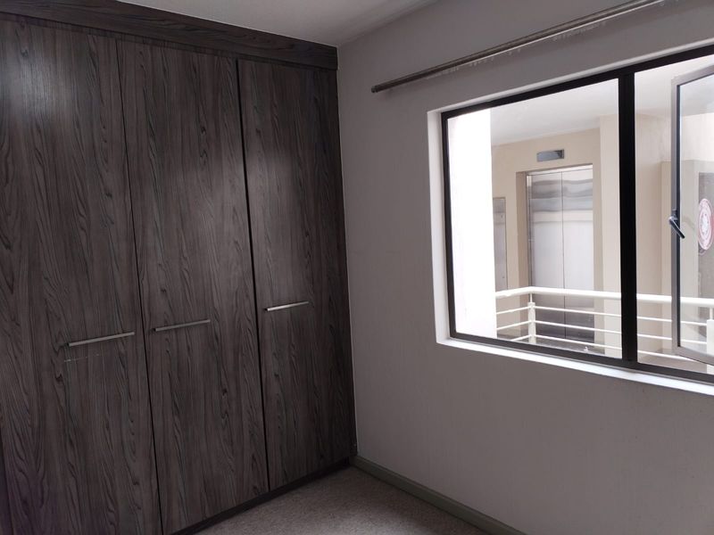 A clean, empty bedroom featuring dark wood-grain built-in cupboards and a window overlooking the apartment building's common corridor and elevator area.