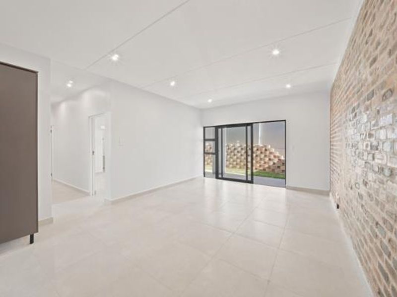 A bright, modern living area featuring a striking exposed brick feature wall, neutral tiled flooring, and large sliding glass doors leading to the exterior.