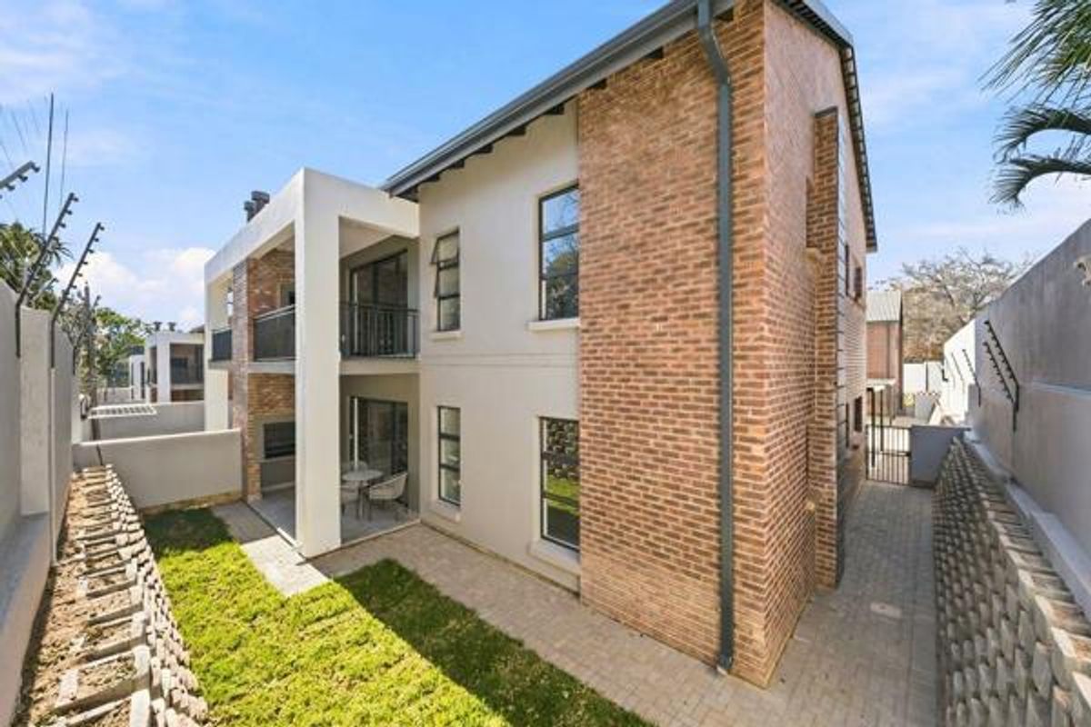 Rear view of a modern apartment building featuring a brick and plaster facade, covered patio, balcony, and a private paved garden area.
