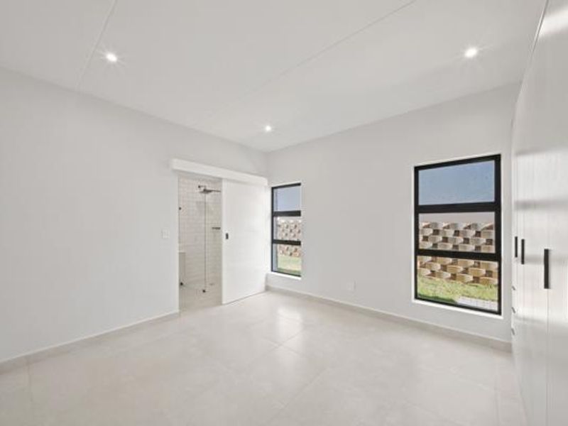 A bright, modern bedroom featuring neutral tiled flooring, built-in cupboards, recessed lighting, and an en-suite bathroom accessed via a sliding door.