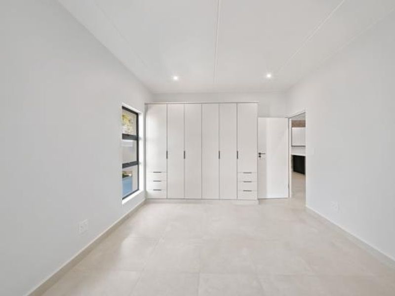 A bright, modern bedroom featuring neutral tiled flooring, ample built-in cupboard storage, and recessed ceiling lighting.