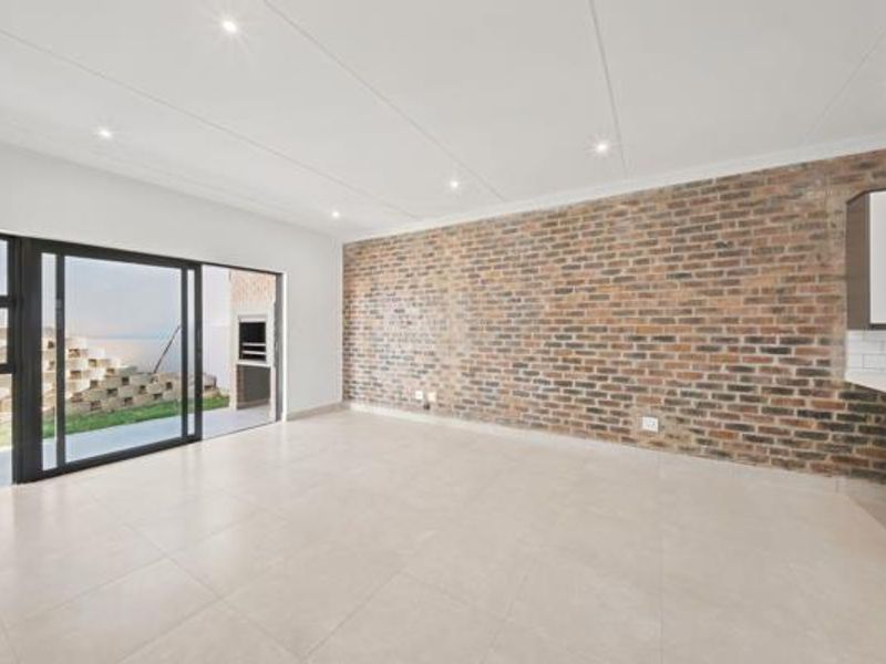 Spacious open-plan living area featuring a striking exposed brick wall, modern tiled flooring, recessed lighting, and seamless access to an outdoor patio with braai.