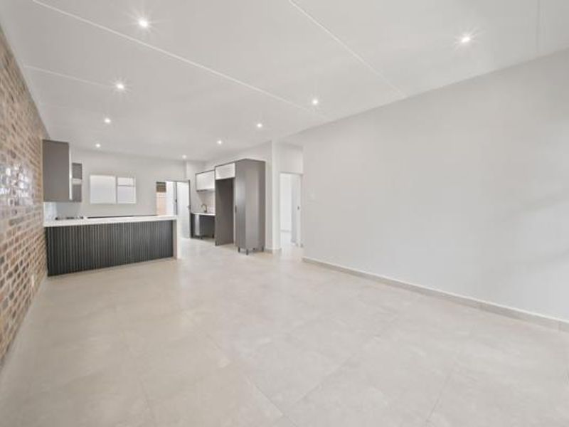 Spacious open-plan living and kitchen area featuring modern tiled flooring, recessed lighting, and a striking exposed brick feature wall.