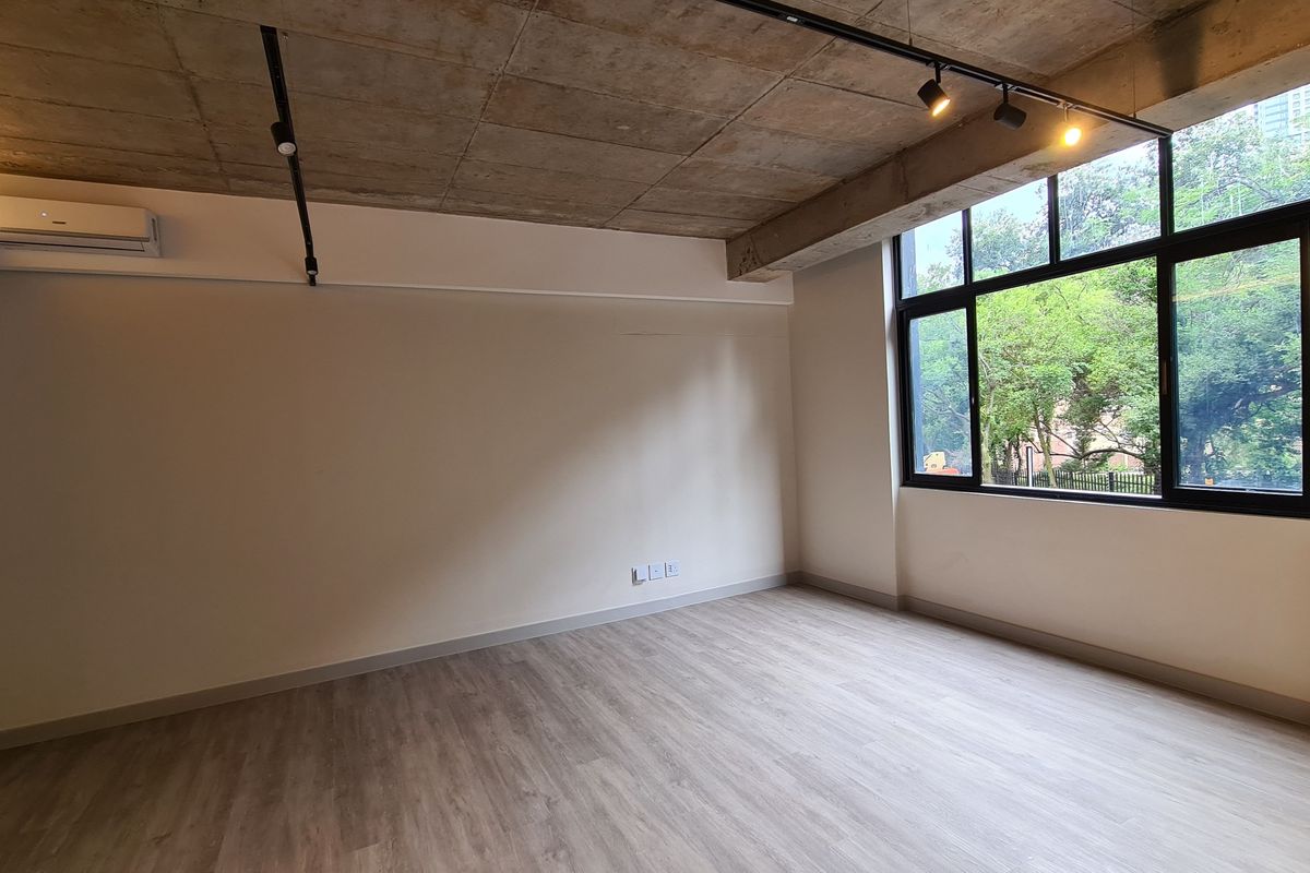 A bright, modern living area featuring an exposed concrete ceiling, track lighting, split-system air conditioning, and large windows with a pleasant green outlook.