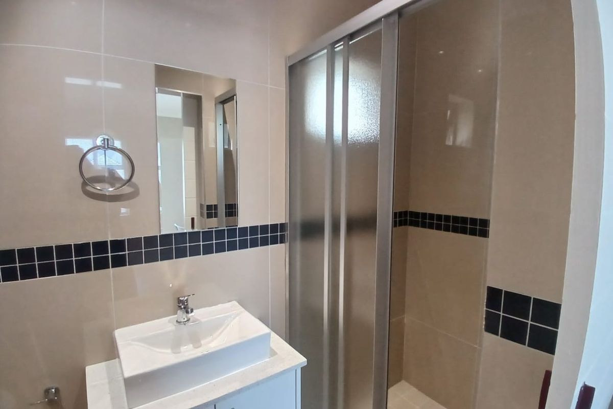 A clean, modern bathroom featuring a vessel sink vanity, wall-mounted mirror, and a glass-enclosed shower with neutral wall tiling and decorative accent borders.