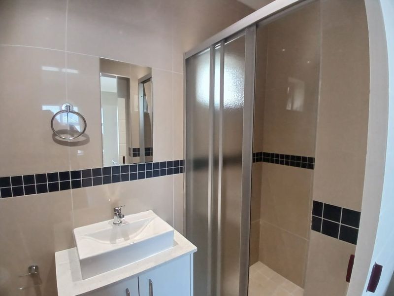A clean, modern bathroom featuring a vessel sink vanity, wall-mounted mirror, and a glass-enclosed shower with neutral wall tiling and decorative accent borders.