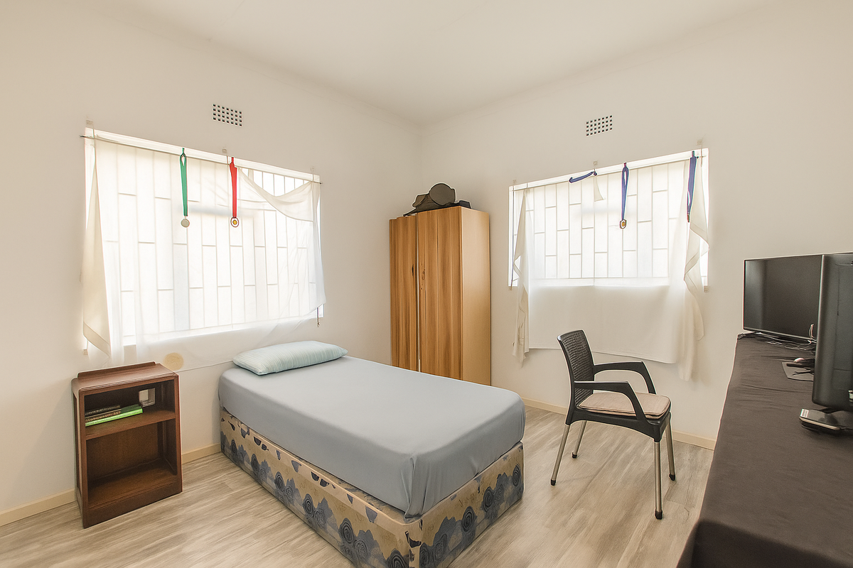 A functional bedroom featuring light-toned wood-look flooring, dual windows with security bars, and neutral wall finishes.