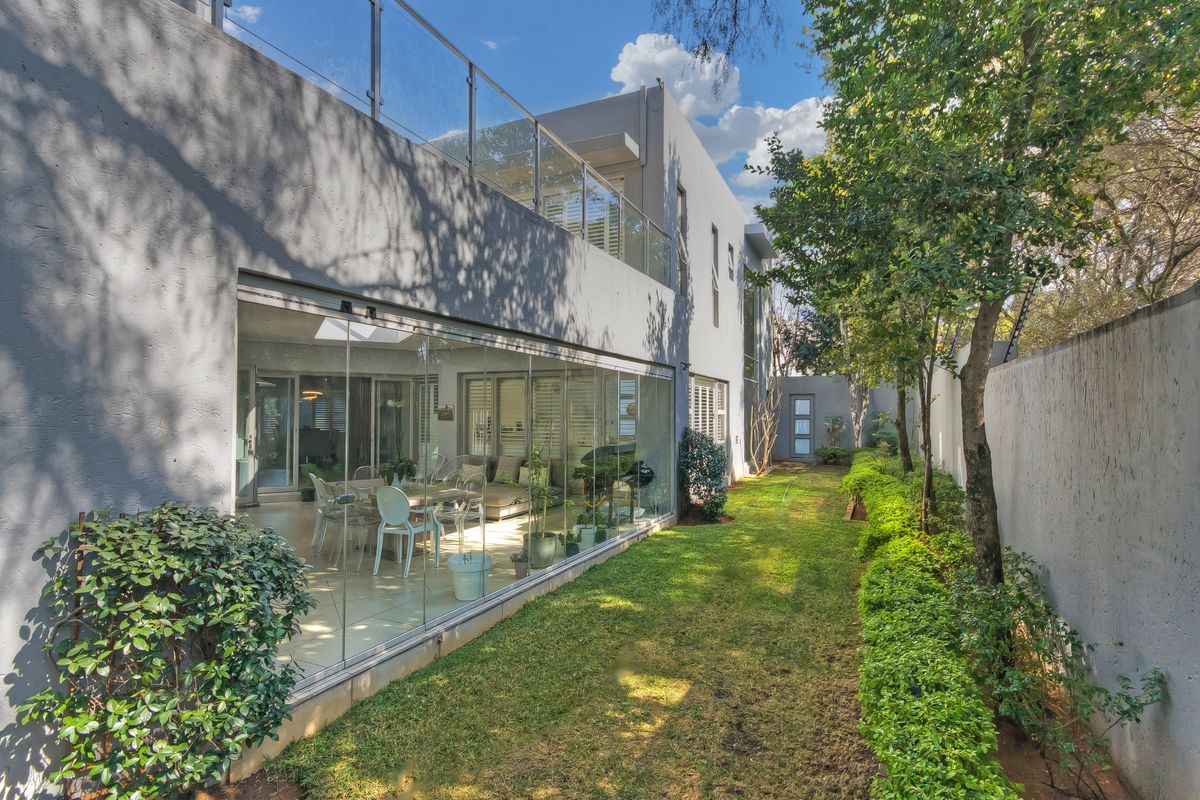 Rear view of a modern cluster home featuring a landscaped garden, glass-enclosed living area, and an upper-level balcony with glass balustrades.