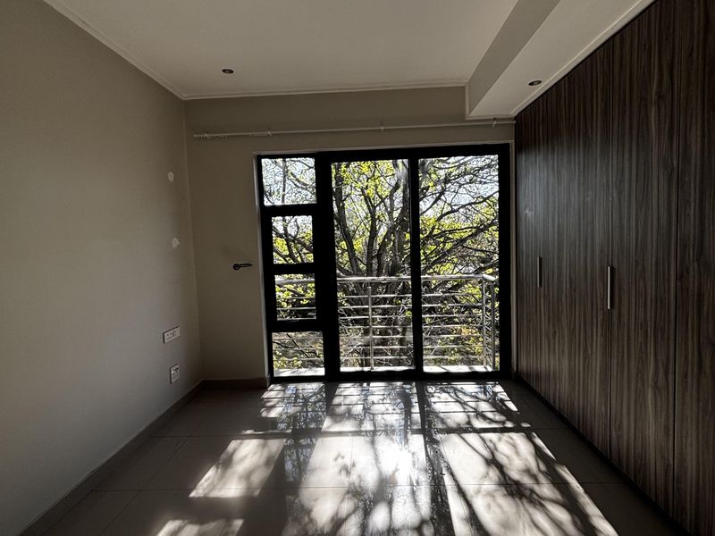 A bright, empty bedroom featuring large built-in wooden wardrobes, tiled flooring, and a glass sliding door leading to a private balcony with tree views.