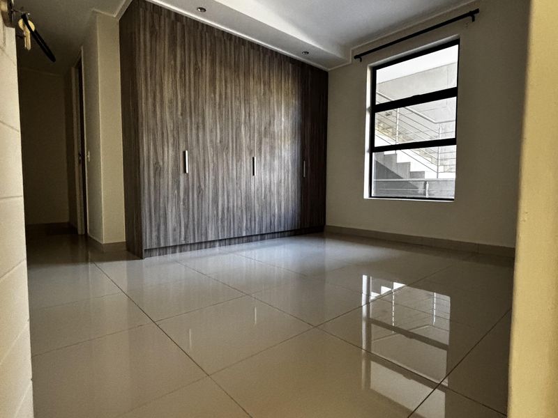A bright, empty bedroom featuring modern large-format floor tiles, a recessed ceiling detail, and built-in floor-to-ceiling wooden cupboards.