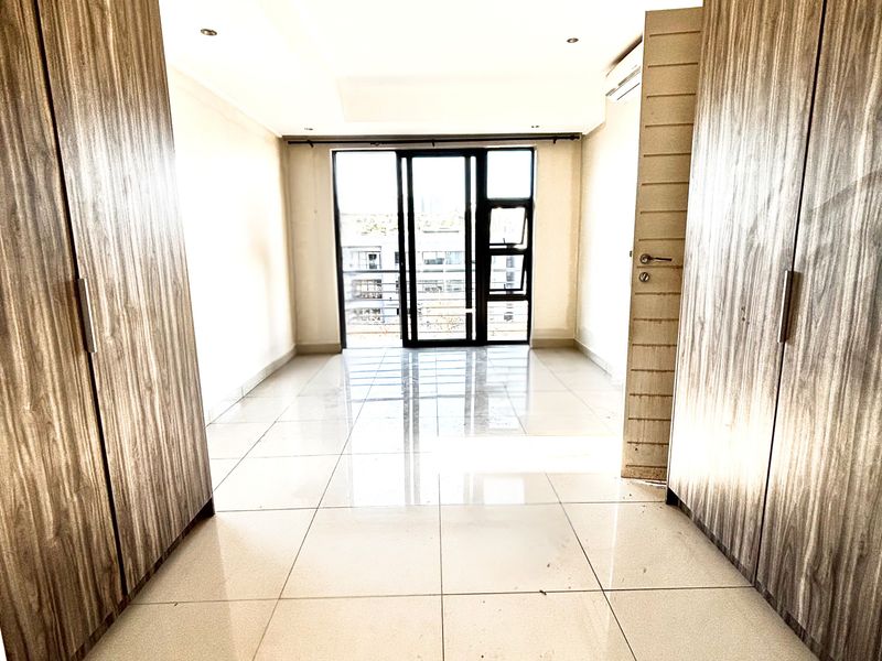 A bright bedroom featuring built-in wooden wardrobes, polished floor tiles, and a glass sliding door leading to a balcony.