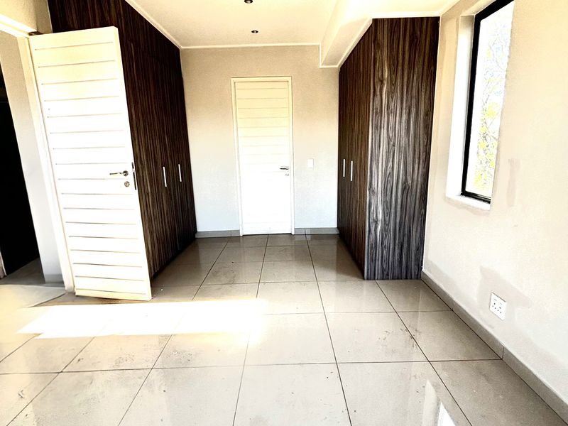 A bright, empty bedroom featuring modern dark wood built-in cupboards, tiled flooring, and recessed ceiling lighting.