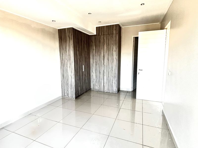 A bright, empty bedroom featuring modern wood-grain built-in cupboards, polished floor tiles, recessed ceiling lighting, and a wall-mounted air conditioning unit.