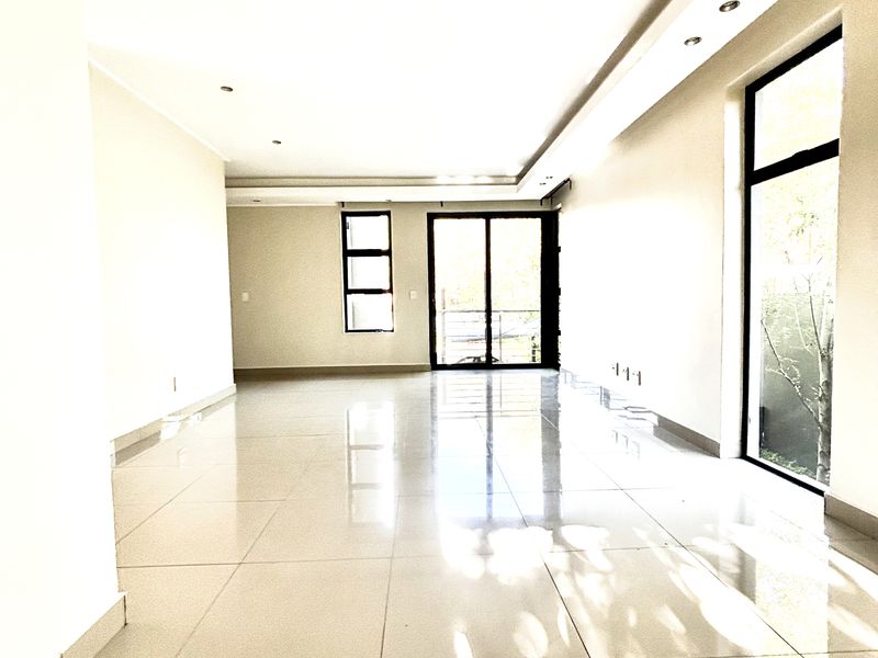 A bright and spacious open-plan living area featuring large format floor tiles, recessed ceiling lighting, and expansive windows providing natural light.