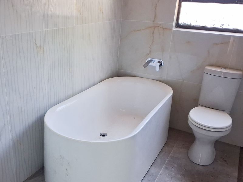 A modern bathroom featuring a freestanding bathtub, wall-mounted mixer, and a standard toilet with neutral wall and floor tiling.