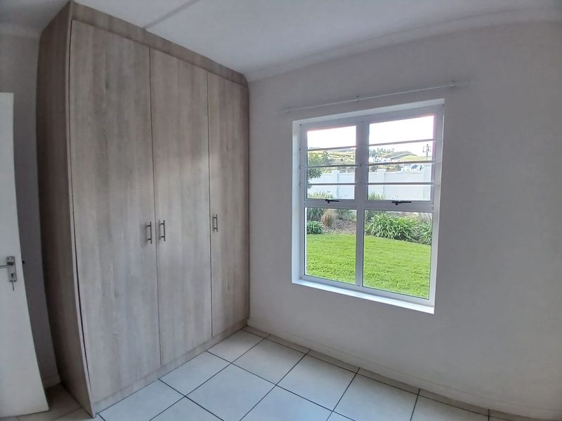 A bright, empty bedroom featuring modern wood-finish built-in cupboards, neutral tiled flooring, and a window with security bars overlooking the garden.