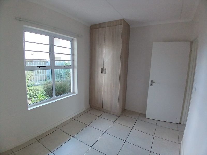 A clean, empty bedroom featuring neutral tiled flooring, a built-in wooden wardrobe, and a window with security bars overlooking the garden.