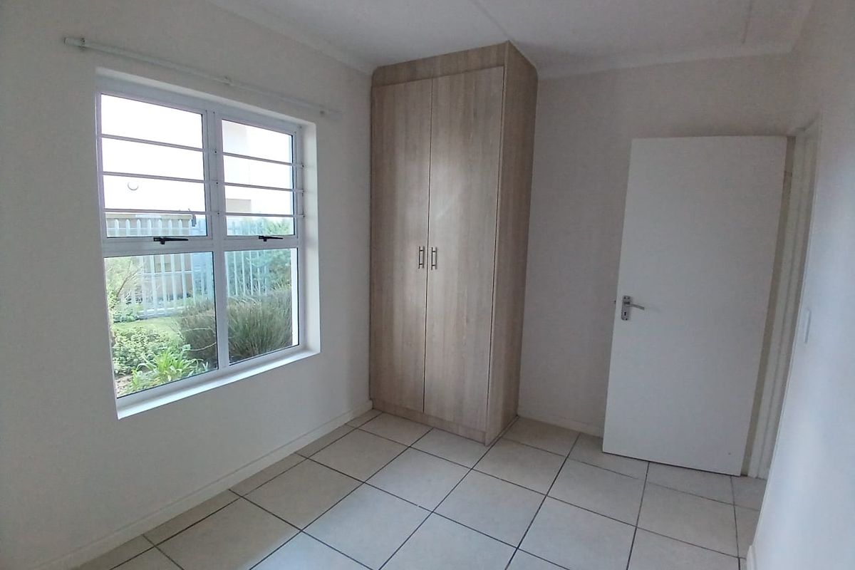 A clean, empty bedroom featuring neutral tiled flooring, a built-in wooden wardrobe, and a window with security bars overlooking the garden.