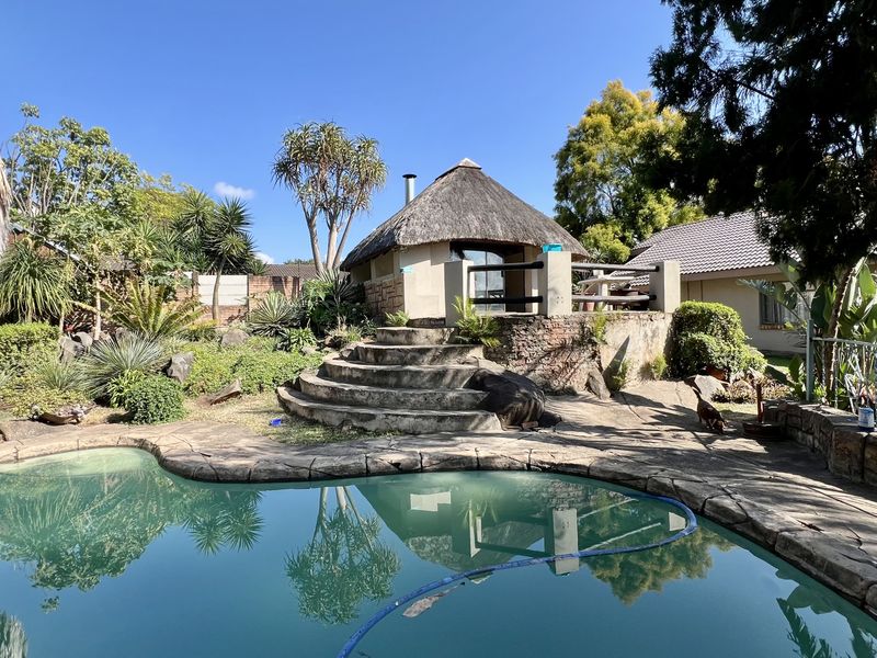 Outdoor pool area featuring a swimming pool, stone paving, and a raised thatched lapa structure set within a landscaped garden.