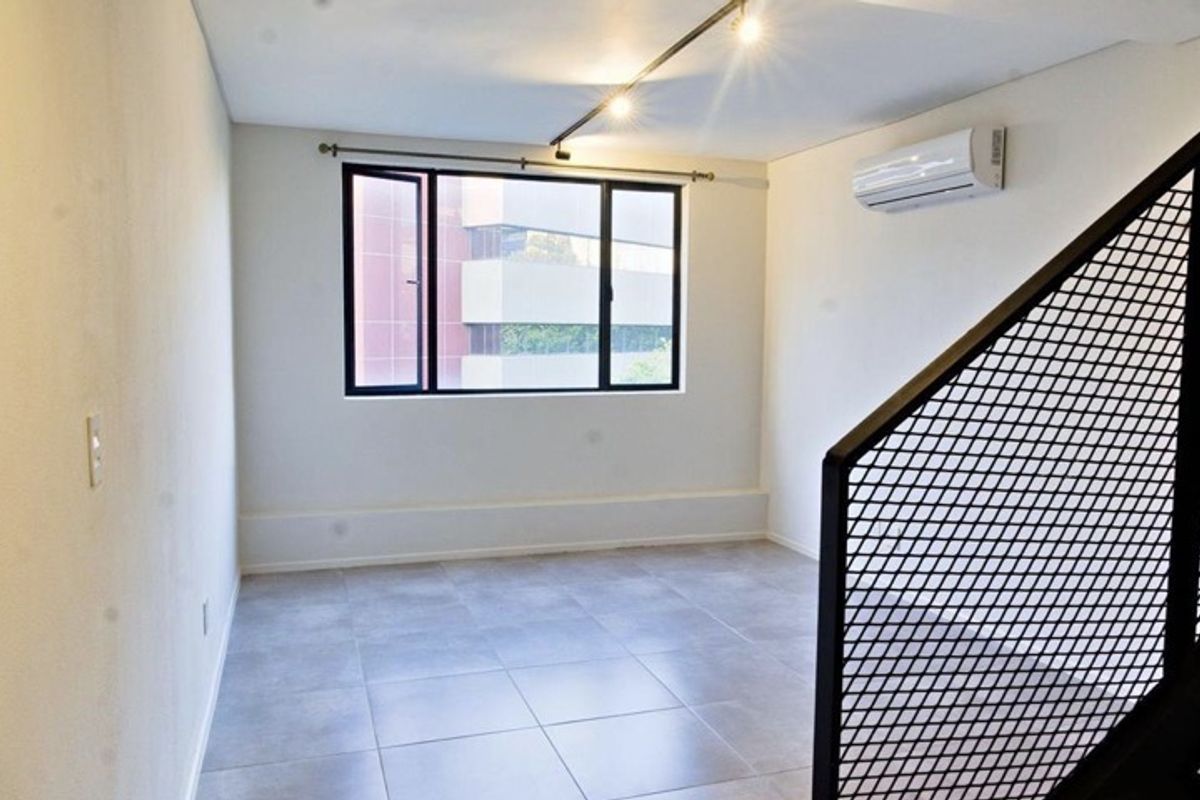 A bright, empty living area featuring neutral tiled flooring, a wall-mounted air conditioning unit, and a modern metal mesh staircase railing.