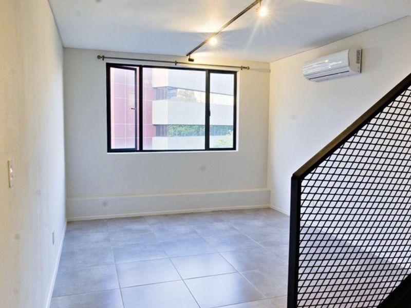 A bright, empty living area featuring neutral tiled flooring, a wall-mounted air conditioning unit, and a modern metal mesh staircase railing.