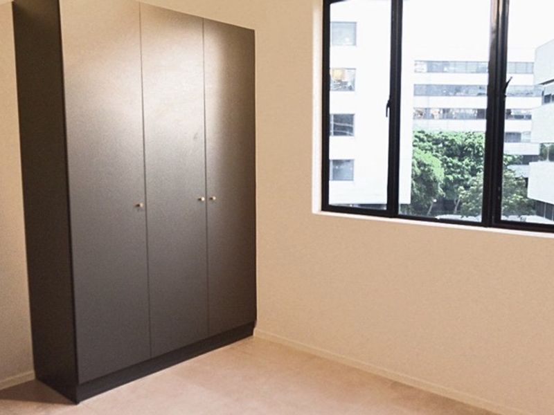 A bright, empty bedroom featuring a modern dark-toned built-in wardrobe and large windows with an urban view.