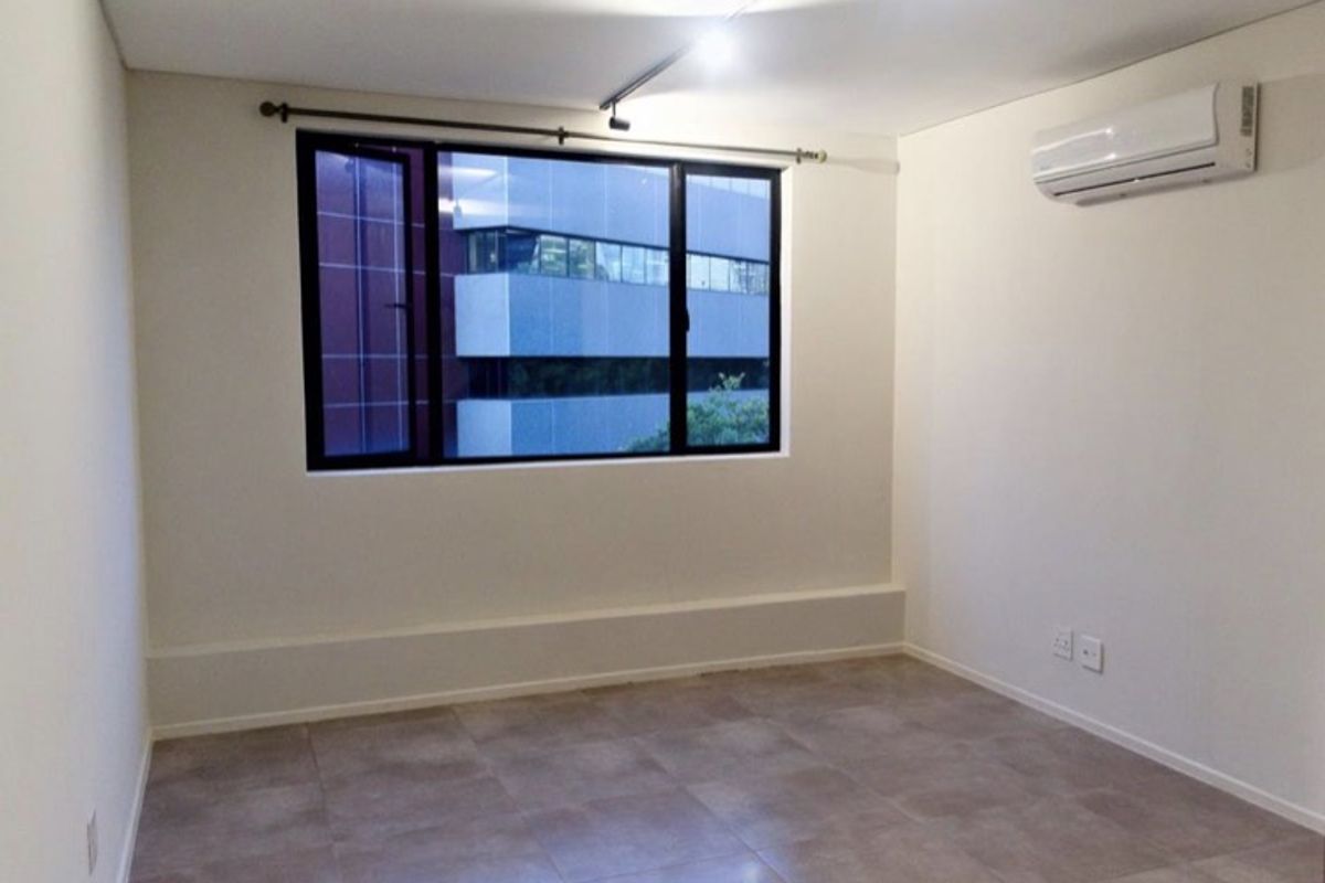 A clean, empty bedroom featuring tiled flooring, a split-system air conditioner, and a large window with a view of the surrounding urban area.
