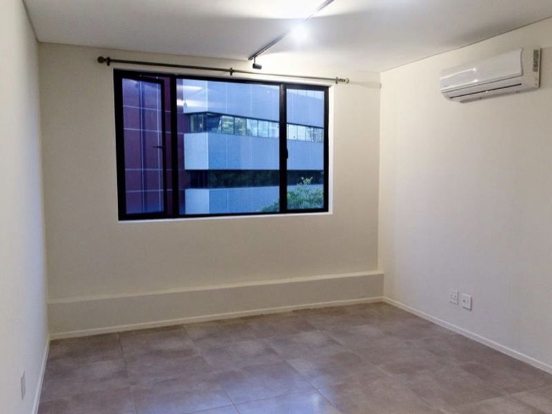 A clean, empty bedroom featuring tiled flooring, a split-system air conditioner, and a large window with a view of the surrounding urban area.