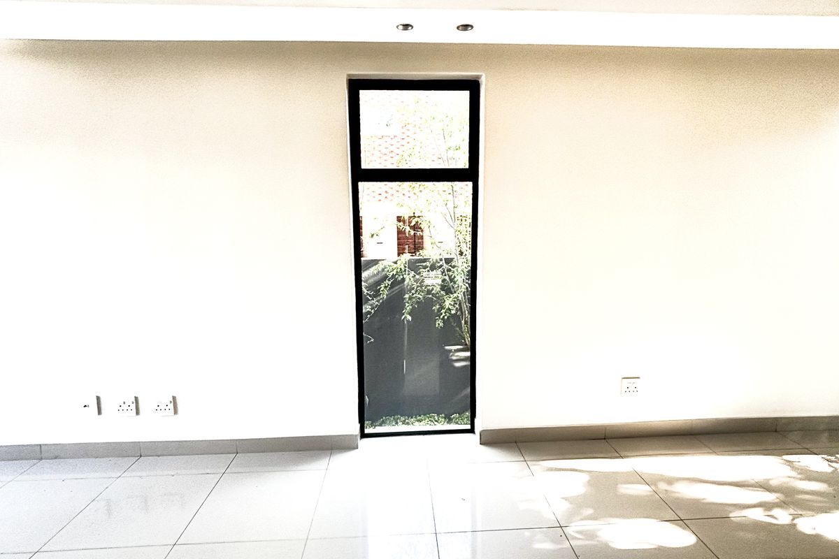 A bright, empty living space featuring polished porcelain floor tiles, recessed ceiling lighting, and a tall, narrow window overlooking a private garden area.