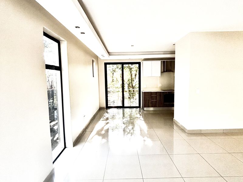 Spacious open-plan living area featuring polished floor tiles, recessed ceiling lighting, and a modern kitchen with built-in cabinetry, leading to a glass-panelled balcony door.