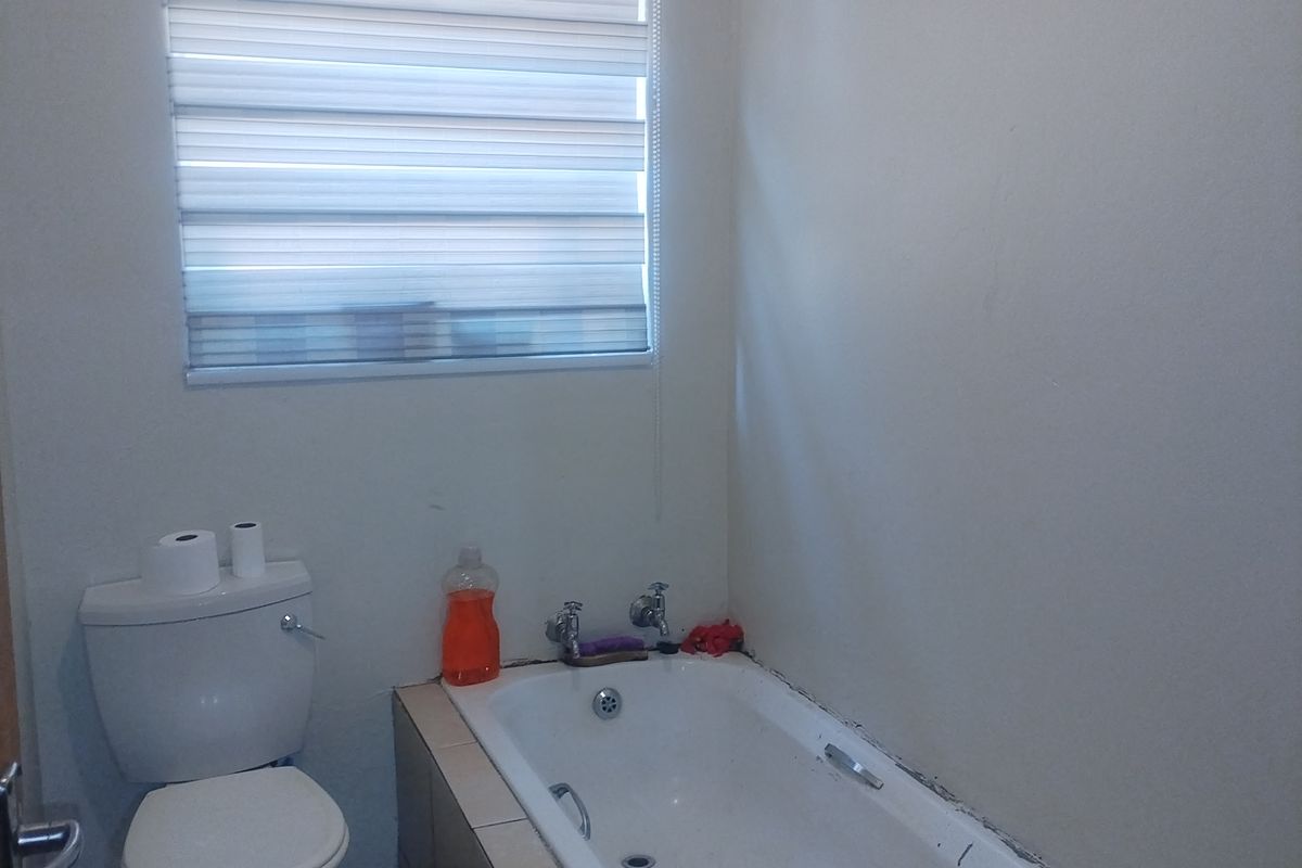 A functional bathroom featuring a built-in bathtub with tiled surround, a standard toilet, and a window with a fitted roller blind.