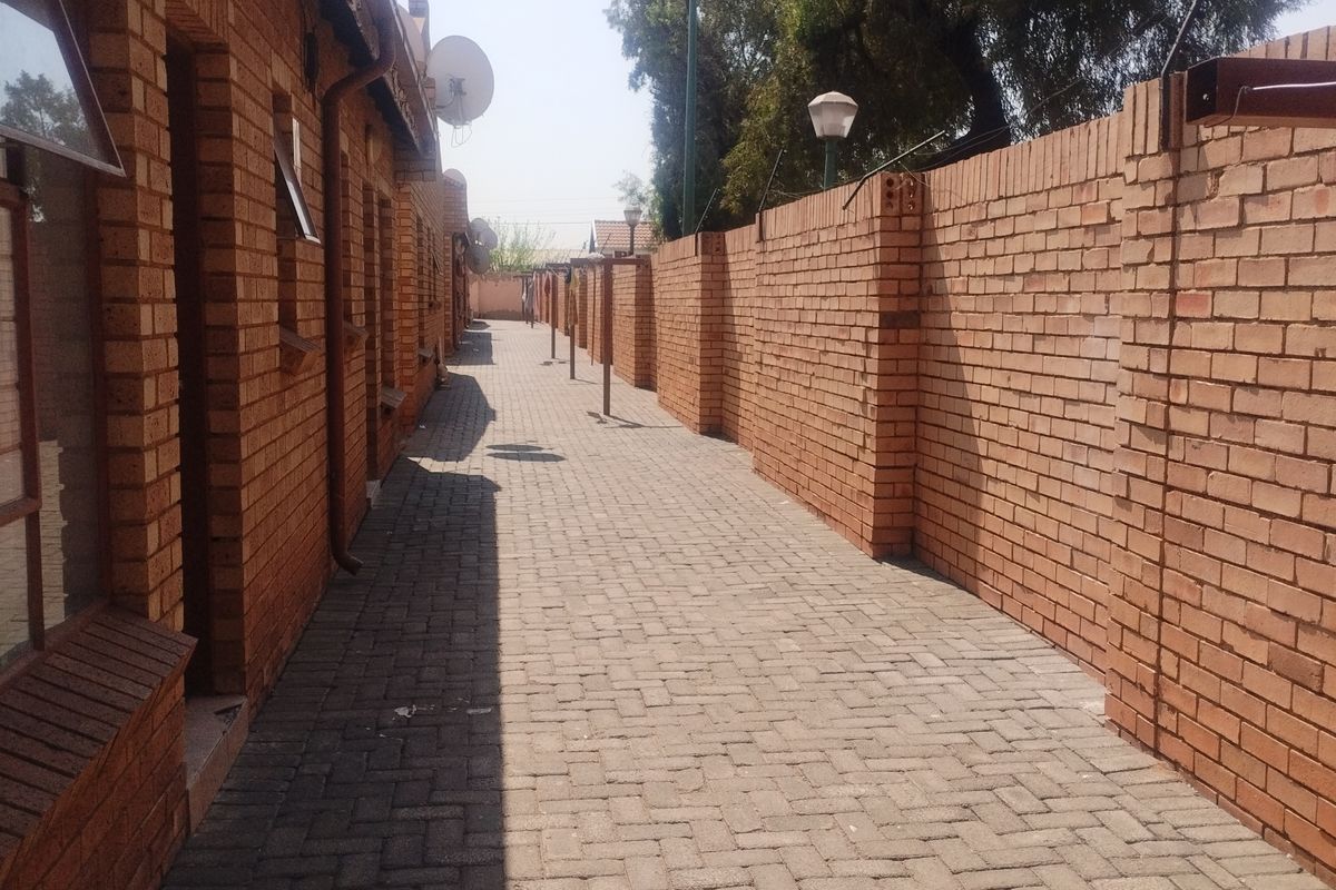 A paved exterior walkway running alongside the apartment building, featuring exposed brick walls and perimeter security fencing.
