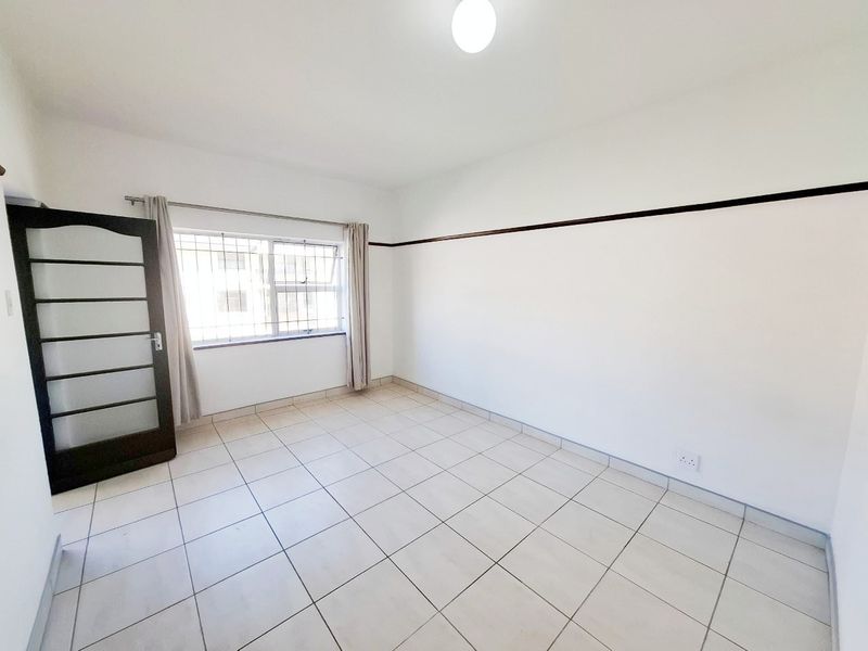 A bright, empty bedroom featuring tiled flooring, a picture rail, and a window fitted with security bars.