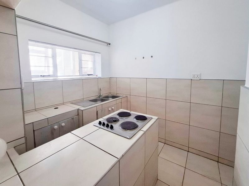 A functional kitchen featuring tiled countertops, built-in cabinetry, a stainless steel sink, and an integrated electric hob with neutral wall tiling.
