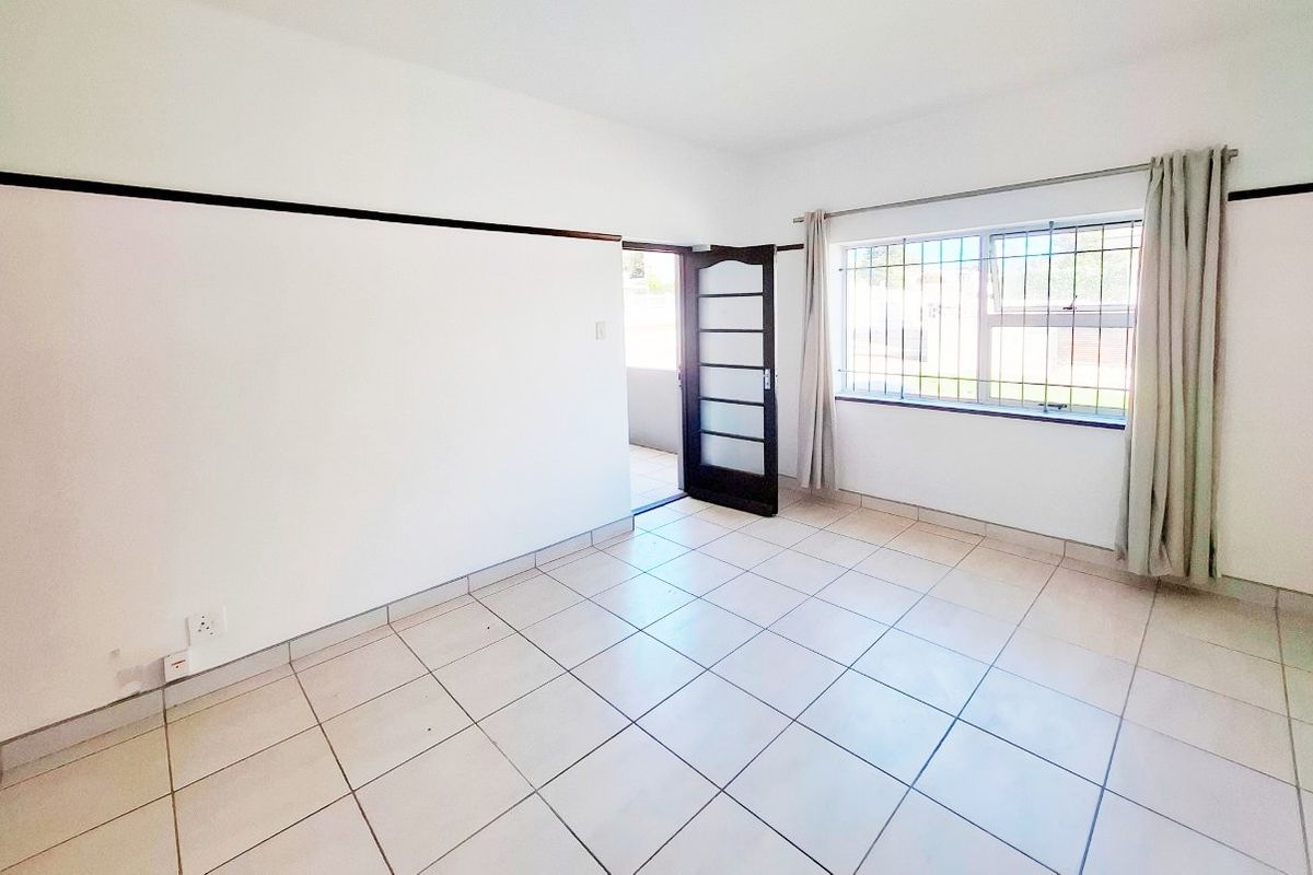 A bright, empty living room featuring neutral tiled flooring, security bars on the window, and a secure external door leading to the outside.