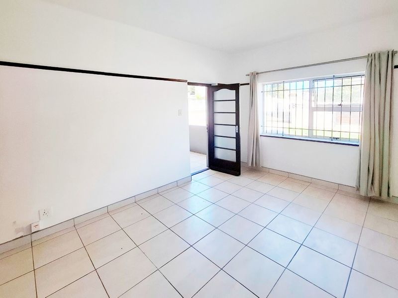 A bright, empty living room featuring neutral tiled flooring, security bars on the window, and a secure external door leading to the outside.
