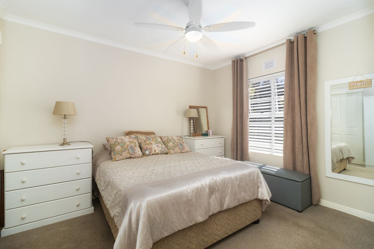 A well-maintained bedroom featuring neutral wall tones, carpeted flooring, a ceiling fan, and a window with fitted blinds and curtains.