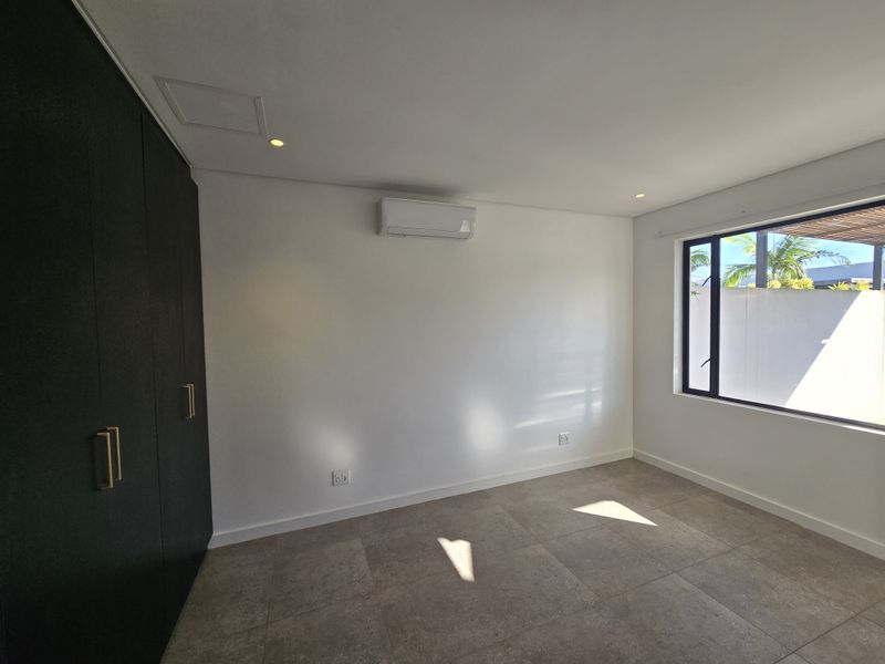 A bright, empty bedroom featuring modern dark built-in cupboards, neutral tiled flooring, and a wall-mounted air conditioning unit.