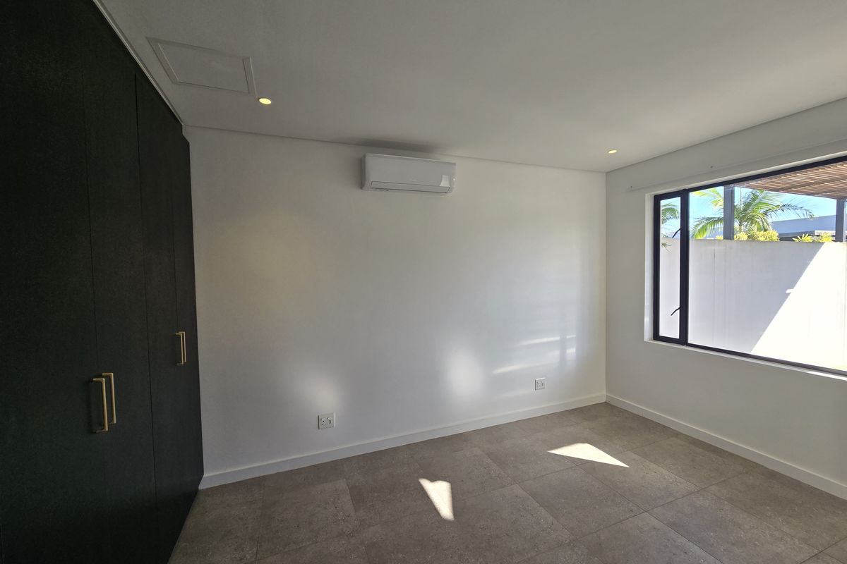 A bright, empty bedroom featuring modern dark built-in cupboards, neutral tiled flooring, and a wall-mounted air conditioning unit.