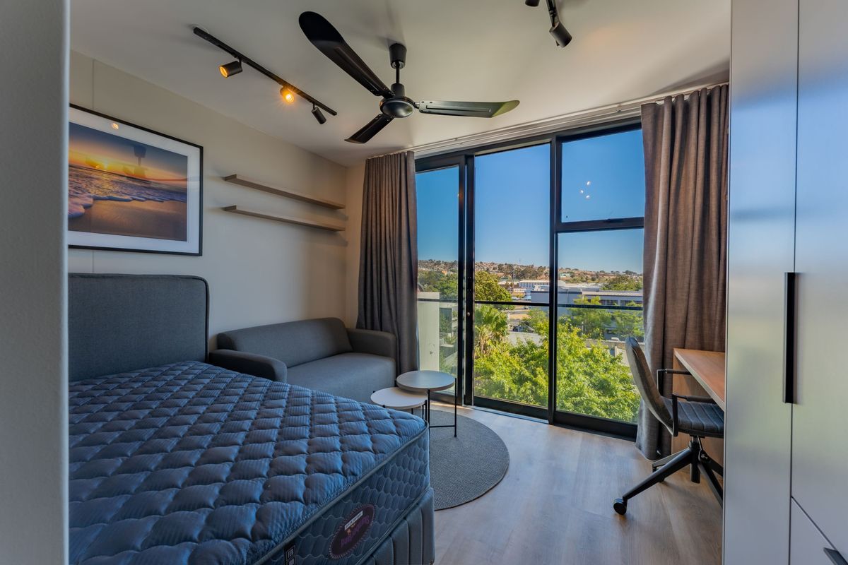 A modern bedroom featuring floor-to-ceiling glass sliding doors, a ceiling fan, track lighting, and built-in shelving, offering pleasant views of the surrounding area.