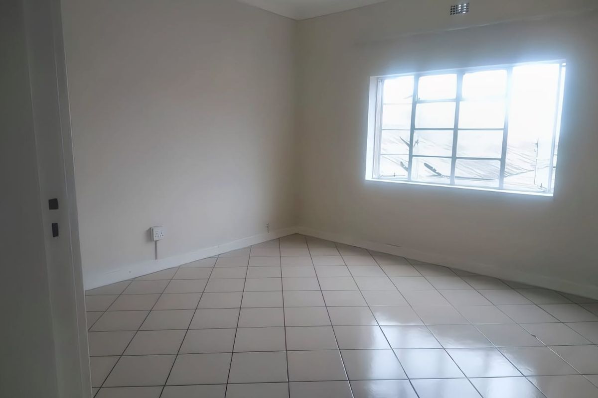 A bright, empty bedroom featuring neutral walls, tiled flooring, and a large steel-framed window providing natural light.