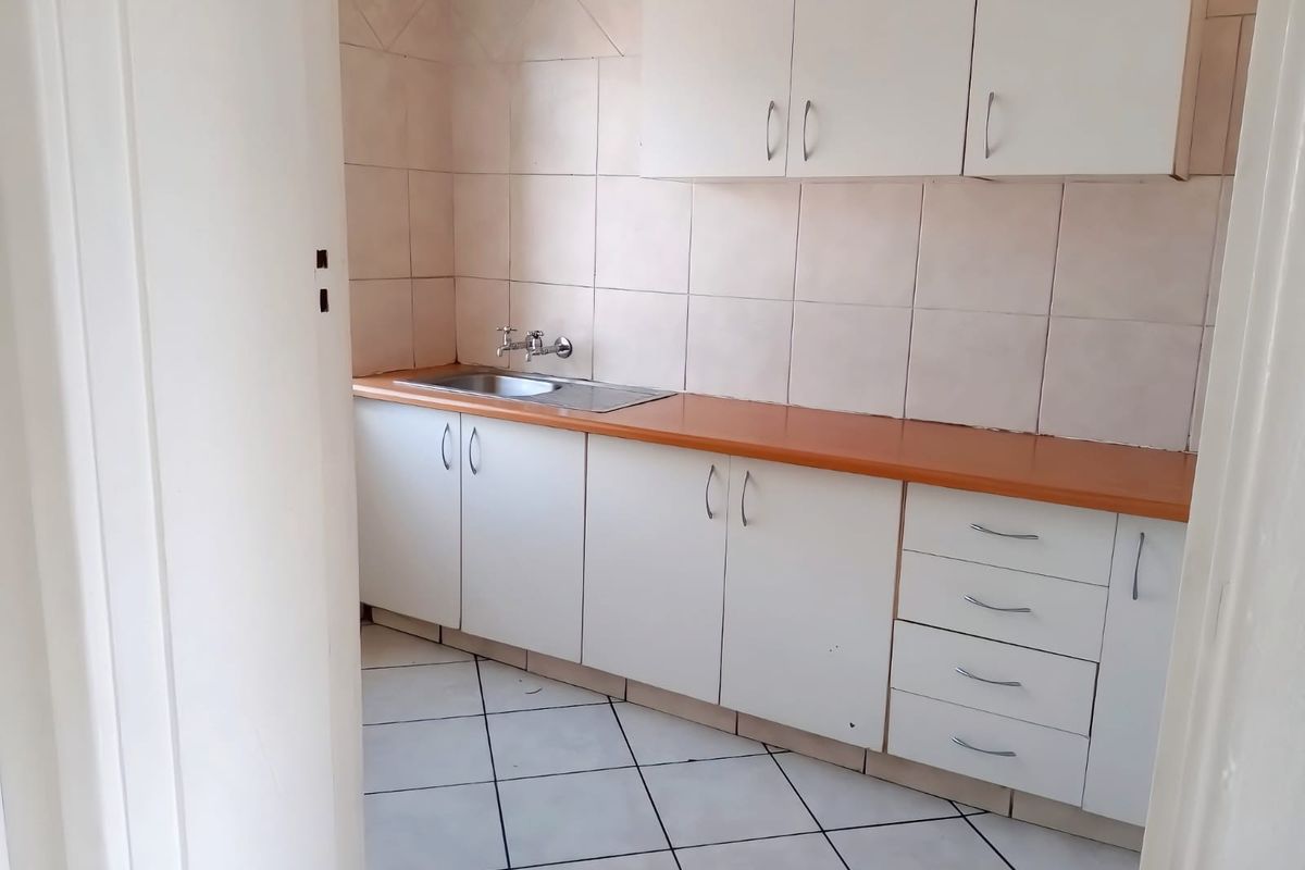 A compact, empty kitchen featuring white built-in cabinetry, a laminate countertop, stainless steel sink, and tiled flooring.