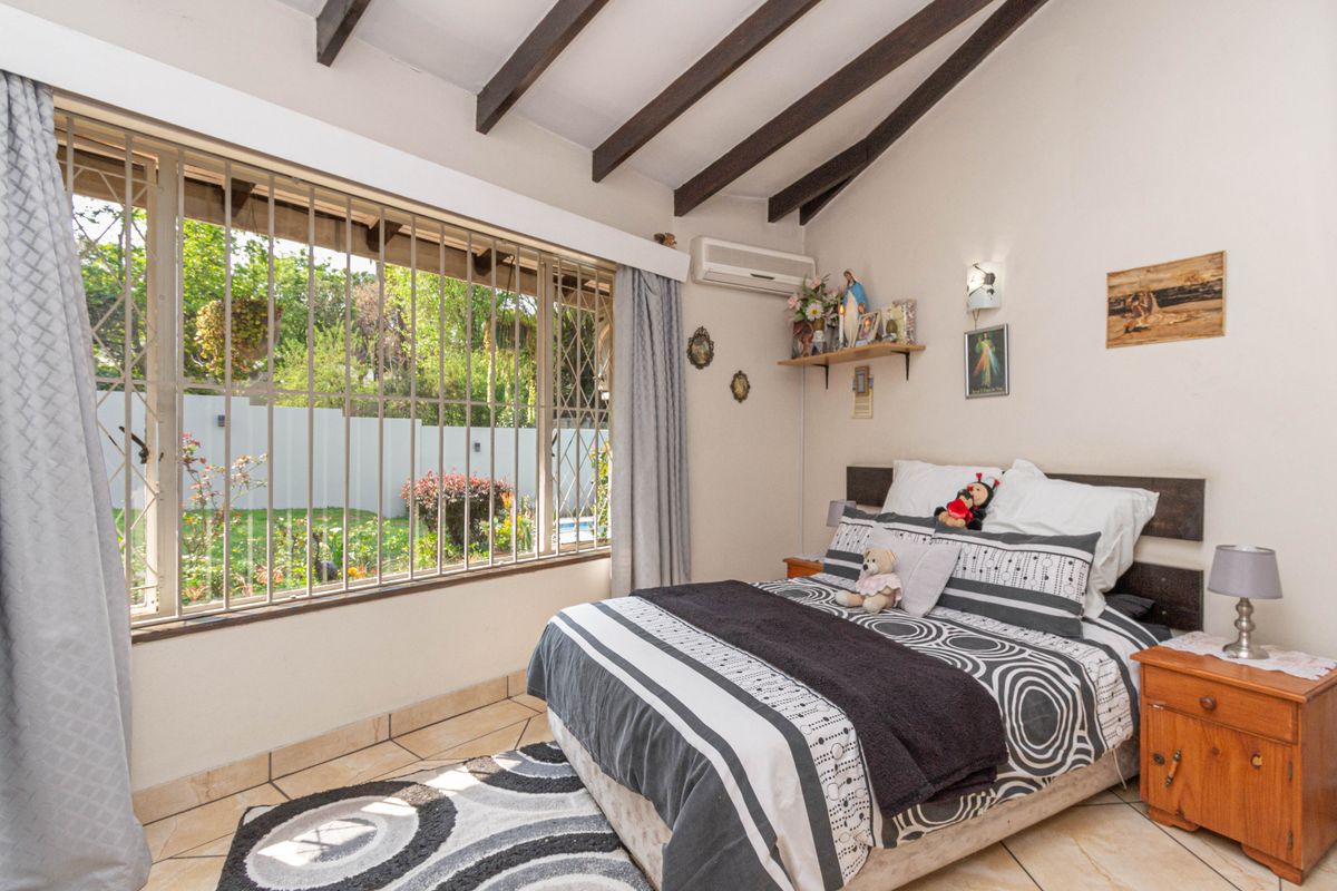 A bedroom featuring exposed timber ceiling beams, tiled flooring, and a large window with security bars overlooking the garden.