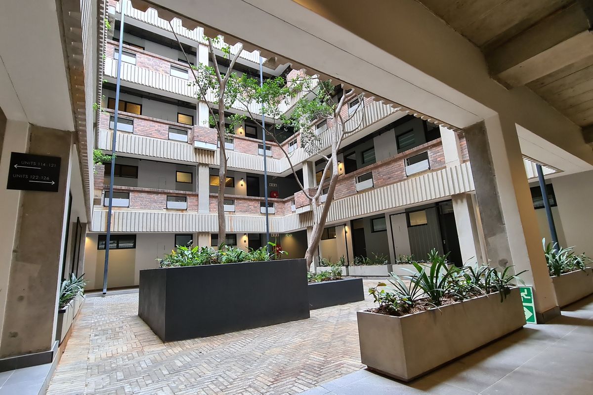 An open-air central courtyard within an apartment complex, featuring brickwork facades, concrete structural elements, and integrated planter boxes with established trees.