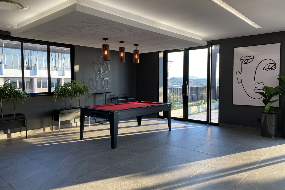 A modern, well-lit entertainment area featuring large windows, tiled flooring, recessed ceiling lighting, and direct access to an outdoor balcony.