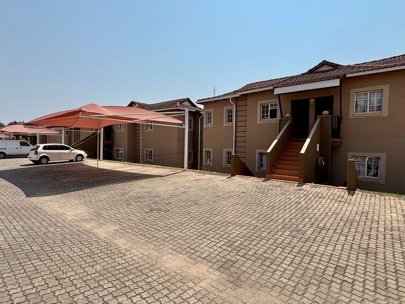 Exterior view of an apartment complex featuring a paved driveway, covered parking bays with shade sails, and multi-level residential units with tiled roofs.